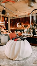 A white, rounded dessert garnished with fresh berries sits on a plate dusted with powdered sugar. The background reveals a cozy restaurant interior with warm lighting, elegant decor, and neatly set tables.
