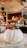 A white, rounded dessert garnished with fresh berries sits on a plate dusted with powdered sugar. The background reveals a cozy restaurant interior with warm lighting, elegant decor, and neatly set tables.