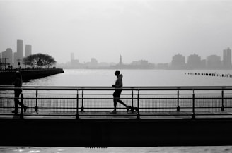 A man is jogging on a bridge with a dog on a leash, set against a backdrop of a city skyline and a large body of water. The silhouette of the jogger and other pedestrians is captured in a monochrome color palette, creating a serene and contemplative atmosphere.