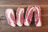 Four slices of raw beef are seasoned with salt and pepper, arranged on a wooden cutting board. The marbling of the meat is prominently visible.