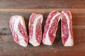 Four slices of raw beef are seasoned with salt and pepper, arranged on a wooden cutting board. The marbling of the meat is prominently visible.
