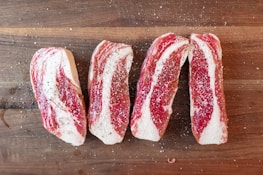 Four slices of raw beef are seasoned with salt and pepper, arranged on a wooden cutting board. The marbling of the meat is prominently visible.