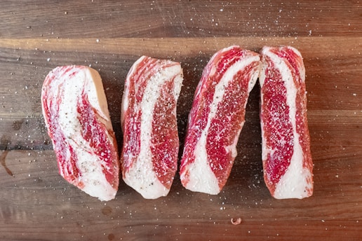 Four slices of raw beef are seasoned with salt and pepper, arranged on a wooden cutting board. The marbling of the meat is prominently visible.