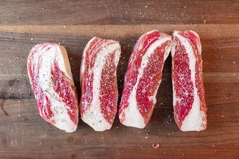 Four slices of raw beef are seasoned with salt and pepper, arranged on a wooden cutting board. The marbling of the meat is prominently visible.