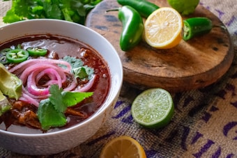 A hearty bowl of rich red stew garnished with sliced jalapenos, pickled red onions, avocado slices, and cilantro. The scene includes a rustic wooden board with fresh ingredients like halved lime and lemon, whole jalapenos, and sprigs of cilantro, all set against a textured fabric background.