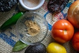 A variety of fresh ingredients are arranged on a textured burlap surface, including a green chili, an onion, ripe red tomatoes, a lemon, an avocado, and a spice dish with ground powder. A bay leaf is also visible, enhancing the rustic and natural vibe.
