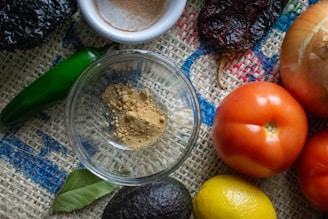 A variety of fresh ingredients are arranged on a textured burlap surface, including a green chili, an onion, ripe red tomatoes, a lemon, an avocado, and a spice dish with ground powder. A bay leaf is also visible, enhancing the rustic and natural vibe.