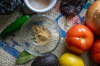 A variety of fresh ingredients are arranged on a textured burlap surface, including a green chili, an onion, ripe red tomatoes, a lemon, an avocado, and a spice dish with ground powder. A bay leaf is also visible, enhancing the rustic and natural vibe.
