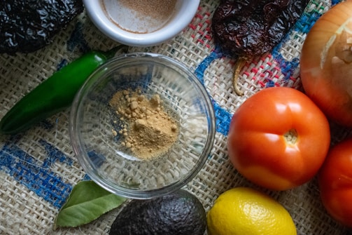 A variety of fresh ingredients are arranged on a textured burlap surface, including a green chili, an onion, ripe red tomatoes, a lemon, an avocado, and a spice dish with ground powder. A bay leaf is also visible, enhancing the rustic and natural vibe.