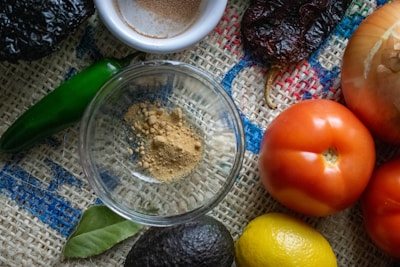 A variety of fresh ingredients are arranged on a textured burlap surface, including a green chili, an onion, ripe red tomatoes, a lemon, an avocado, and a spice dish with ground powder. A bay leaf is also visible, enhancing the rustic and natural vibe.