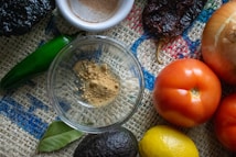 A variety of fresh ingredients are arranged on a textured burlap surface, including a green chili, an onion, ripe red tomatoes, a lemon, an avocado, and a spice dish with ground powder. A bay leaf is also visible, enhancing the rustic and natural vibe.