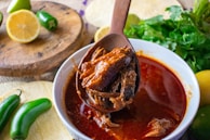 A bowl of rich, red stew with shredded meat, likely birria, is being served with a wooden spoon. Surrounding the bowl are fresh ingredients including lime, jalapeños, and cilantro, which add a vibrant touch to the rustic setting.