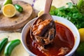 A bowl of rich, red stew with shredded meat, likely birria, is being served with a wooden spoon. Surrounding the bowl are fresh ingredients including lime, jalapeños, and cilantro, which add a vibrant touch to the rustic setting.