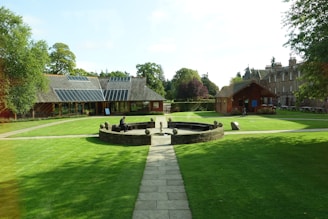 A well-maintained courtyard features a circular stone fountain surrounded by a grassy lawn. Paths intersect at the center, leading to buildings with rustic architectural designs. One structure has solar panels on the roof, and a person is seated near the fountain. The environment is serene, with trees in the background under a clear sky.