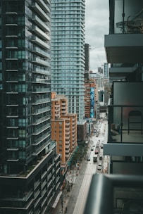 An urban landscape with a street flanked by tall modern buildings. These structures feature large windows and balconies, and the street below is lined with parked cars and small trees. The area appears bustling yet orderly, with people and vehicles visible in the distance. The sky is overcast, casting a muted light over the scene.