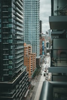 An urban landscape with a street flanked by tall modern buildings. These structures feature large windows and balconies, and the street below is lined with parked cars and small trees. The area appears bustling yet orderly, with people and vehicles visible in the distance. The sky is overcast, casting a muted light over the scene.