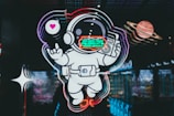A neon-style illustration of an astronaut floating in space, holding a retro radio with a speech bubble displaying a heart. Beside the astronaut, there is a depiction of a planet with rings, resembling Saturn. The background features abstract, colorful reflections, enhancing the futuristic and psychedelic theme.