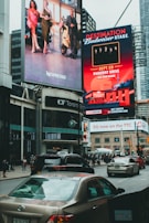 Large, vibrant billboards displaying advertisements and promotions are mounted on the sides of a tall building. Below, a busy urban street is filled with cars and pedestrians, indicating a bustling city atmosphere.