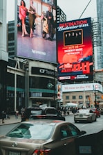 Large, vibrant billboards displaying advertisements and promotions are mounted on the sides of a tall building. Below, a busy urban street is filled with cars and pedestrians, indicating a bustling city atmosphere.