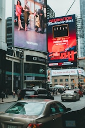 Large, vibrant billboards displaying advertisements and promotions are mounted on the sides of a tall building. Below, a busy urban street is filled with cars and pedestrians, indicating a bustling city atmosphere.