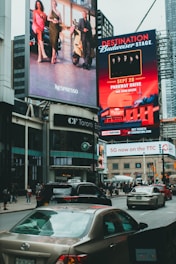Large, vibrant billboards displaying advertisements and promotions are mounted on the sides of a tall building. Below, a busy urban street is filled with cars and pedestrians, indicating a bustling city atmosphere.