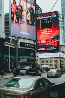 Large, vibrant billboards displaying advertisements and promotions are mounted on the sides of a tall building. Below, a busy urban street is filled with cars and pedestrians, indicating a bustling city atmosphere.