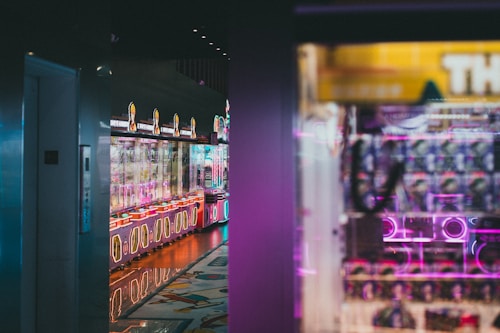 A dimly lit arcade features colorful claw machines with neon lights. Reflective surfaces enhance the glowing ambiance in the corridor.