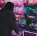 A person is playing a claw machine filled with colorful plush toys. The machine is illuminated by bright purple and pink neon lights, creating a playful atmosphere. Various stuffed animals, including a green toy being picked up by the claw, are visible inside.