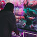 A person is playing a claw machine filled with colorful plush toys. The machine is illuminated by bright purple and pink neon lights, creating a playful atmosphere. Various stuffed animals, including a green toy being picked up by the claw, are visible inside.