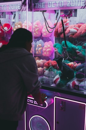 A person is playing a claw machine filled with colorful plush toys. The machine is illuminated by bright purple and pink neon lights, creating a playful atmosphere. Various stuffed animals, including a green toy being picked up by the claw, are visible inside.