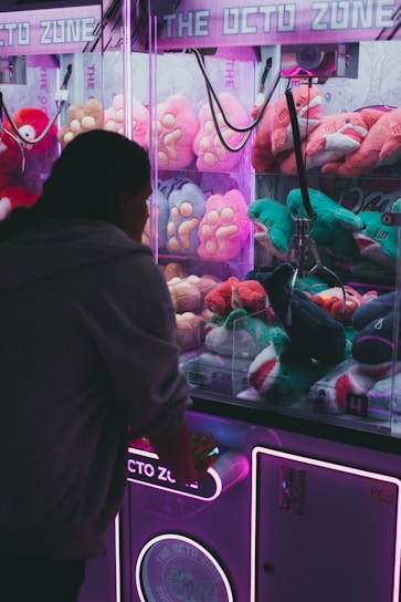 A person is playing a claw machine filled with colorful plush toys. The machine is illuminated by bright purple and pink neon lights, creating a playful atmosphere. Various stuffed animals, including a green toy being picked up by the claw, are visible inside.