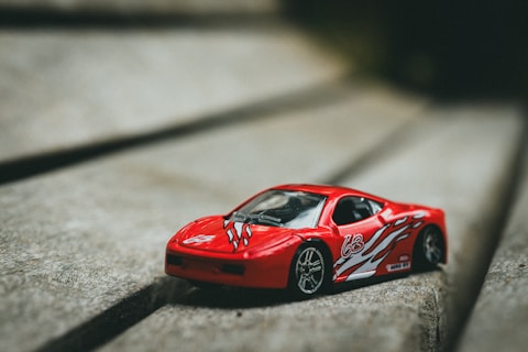 A small red toy car with white and black decals is placed on a textured wooden surface. The car features a sleek design, resembling a sports car, with the number 63 on its side and flame-like decorations.