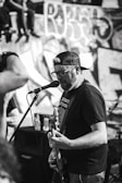 A man in a backwards baseball cap and glasses is passionately singing and playing an electric guitar on stage. He is facing a microphone and appears to be performing in front of a graffiti-covered wall. There are other people visible in the background and foreground, contributing to a lively atmosphere.