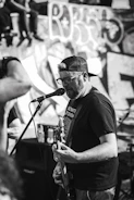 A man in a backwards baseball cap and glasses is passionately singing and playing an electric guitar on stage. He is facing a microphone and appears to be performing in front of a graffiti-covered wall. There are other people visible in the background and foreground, contributing to a lively atmosphere.