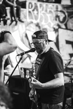 A man in a backwards baseball cap and glasses is passionately singing and playing an electric guitar on stage. He is facing a microphone and appears to be performing in front of a graffiti-covered wall. There are other people visible in the background and foreground, contributing to a lively atmosphere.