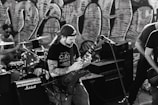 A black and white image capturing a band performing. A guitarist with long hair and a backwards cap plays passionately, surrounded by musical equipment including amplifiers and a drum set. Graffiti art covers the wall in the background, adding an urban vibe to the scene.