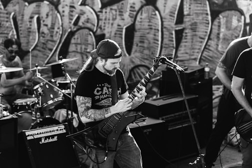 A black and white image capturing a band performing. A guitarist with long hair and a backwards cap plays passionately, surrounded by musical equipment including amplifiers and a drum set. Graffiti art covers the wall in the background, adding an urban vibe to the scene.
