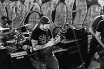 A black and white image capturing a band performing. A guitarist with long hair and a backwards cap plays passionately, surrounded by musical equipment including amplifiers and a drum set. Graffiti art covers the wall in the background, adding an urban vibe to the scene.