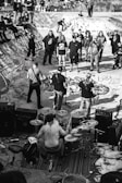 A black and white image depicting a live band performance in a skatepark. The drummer is shirtless and surrounded by his drum set, playing energetically. Several people are gathered around, some standing on the skatepark edges. The skatepark is covered in graffiti, adding to the urban and edgy atmosphere.