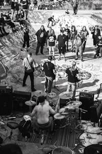 A black and white image depicting a live band performance in a skatepark. The drummer is shirtless and surrounded by his drum set, playing energetically. Several people are gathered around, some standing on the skatepark edges. The skatepark is covered in graffiti, adding to the urban and edgy atmosphere.