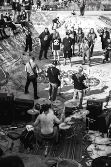A black and white image depicting a live band performance in a skatepark. The drummer is shirtless and surrounded by his drum set, playing energetically. Several people are gathered around, some standing on the skatepark edges. The skatepark is covered in graffiti, adding to the urban and edgy atmosphere.
