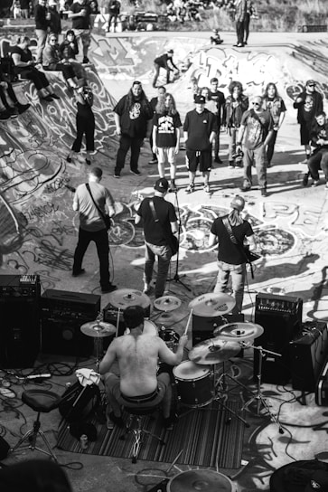 A black and white image depicting a live band performance in a skatepark. The drummer is shirtless and surrounded by his drum set, playing energetically. Several people are gathered around, some standing on the skatepark edges. The skatepark is covered in graffiti, adding to the urban and edgy atmosphere.