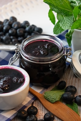 A jar and a bowl filled with dark berry jam are placed on a checkered cloth with fresh berries scattered around. A wooden spoon lies nearby, along with a sprig of fresh mint.