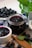 A jar and a bowl filled with dark berry jam are placed on a checkered cloth with fresh berries scattered around. A wooden spoon lies nearby, along with a sprig of fresh mint.
