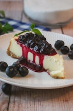 A slice of cheesecake topped with a thick layer of blueberry sauce is served on a white plate. Fresh blueberries are scattered around the plate, and a sprig of mint adds a touch of green. The cheesecake is creamy with a graham cracker crust, and the berry sauce drips invitingly down the side.