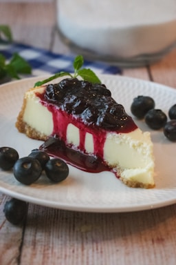 A slice of cheesecake topped with a thick layer of blueberry sauce is served on a white plate. Fresh blueberries are scattered around the plate, and a sprig of mint adds a touch of green. The cheesecake is creamy with a graham cracker crust, and the berry sauce drips invitingly down the side.