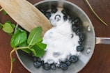 A saucepan containing blueberries topped with sugar is placed on a wooden surface. A wooden spatula is partially submerged in the sugar, and fresh mint leaves are positioned beside the pan.