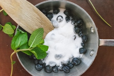 A saucepan containing blueberries topped with sugar is placed on a wooden surface. A wooden spatula is partially submerged in the sugar, and fresh mint leaves are positioned beside the pan.