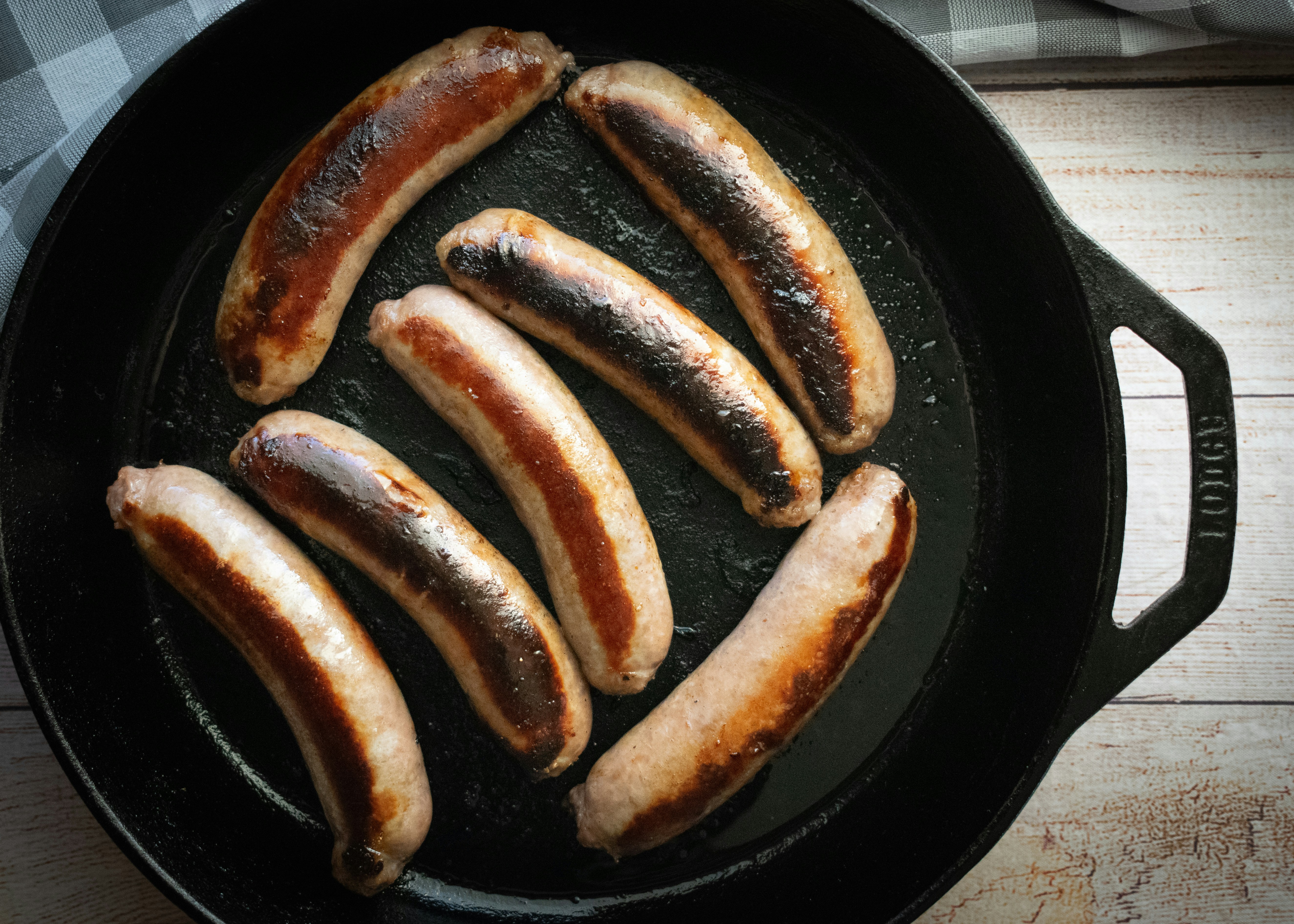 five sausages are cooking in a cast iron skillet, From above, a Lodge cast iron pan showcases perfectly cooked bratwurst, their golden-brown exterior hinting at the savory experience beneath. The arrangement provides a direct glimpse into culinary craftsmanship, capturing the essence of traditional grilling in quality cookware.
