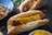 A grilled sausage in a bun topped with yellow mustard, alongside another sausage with sauerkraut in the background. A pan with fried onions and a dish with more mustard are present, suggesting a meal preparation setting.